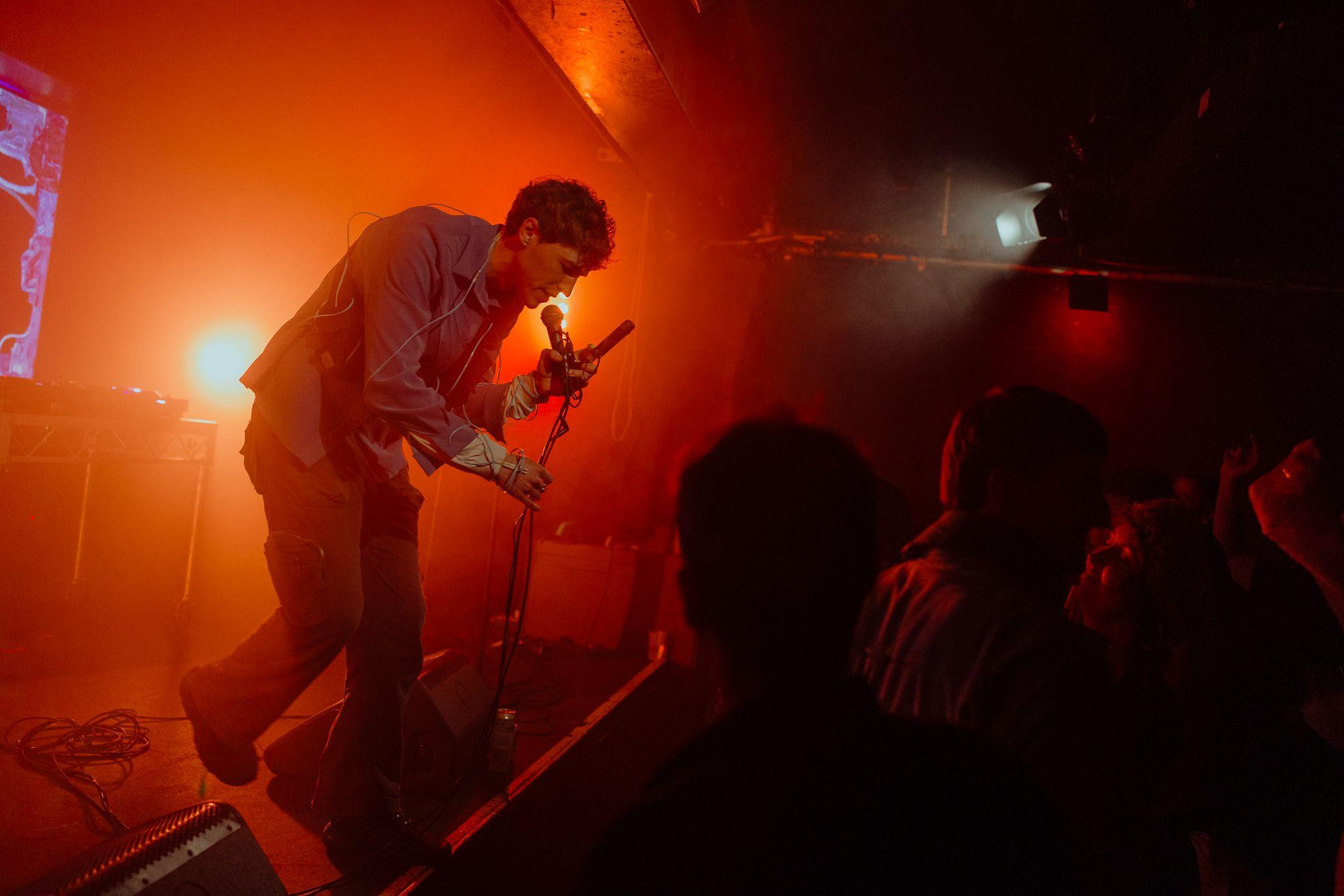 A performer leans into a microphone, towards a crowd, from a stage bathed in orange haze