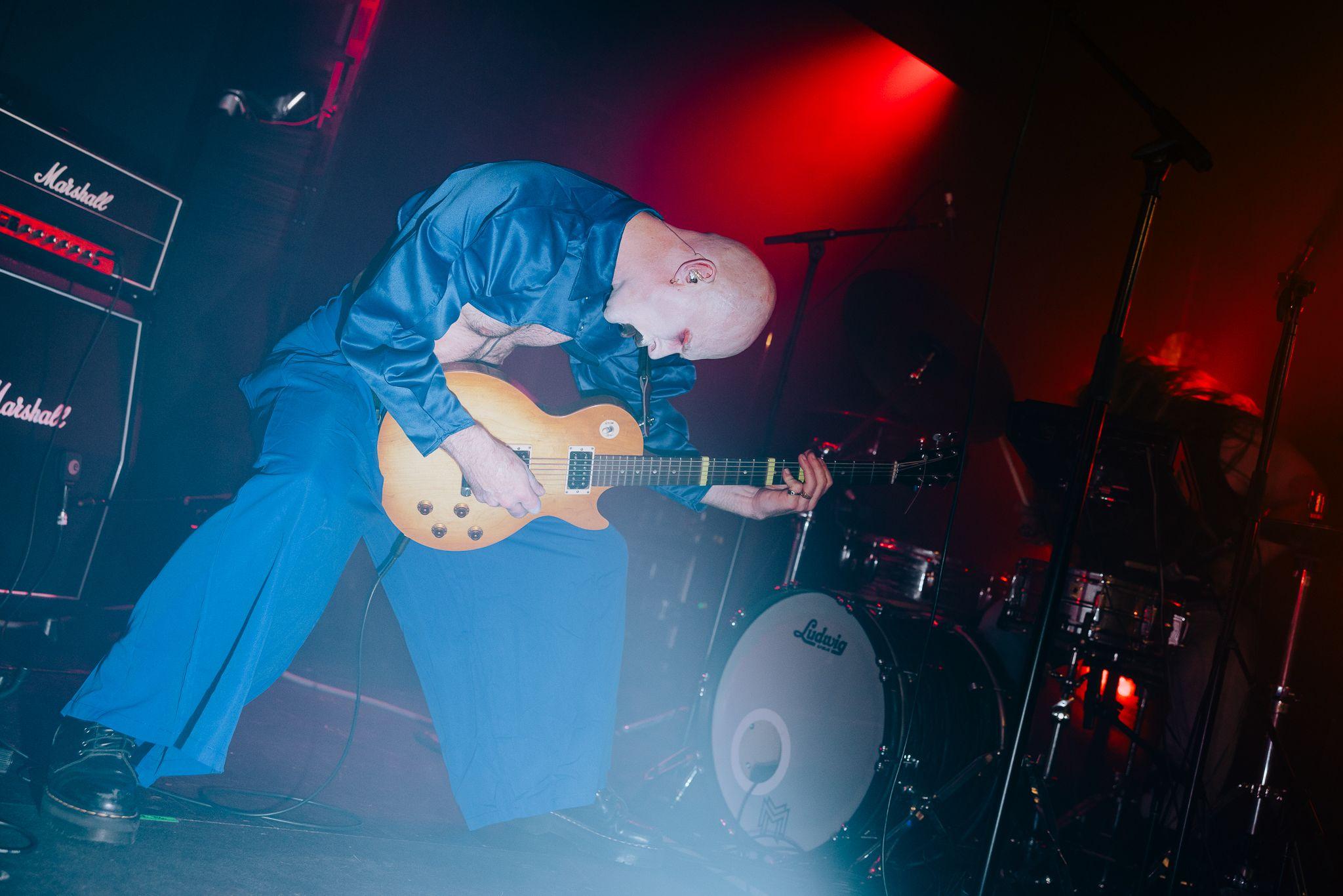 Man on-stage playing guitar, shrouded in shadow a drummer plays the drums.