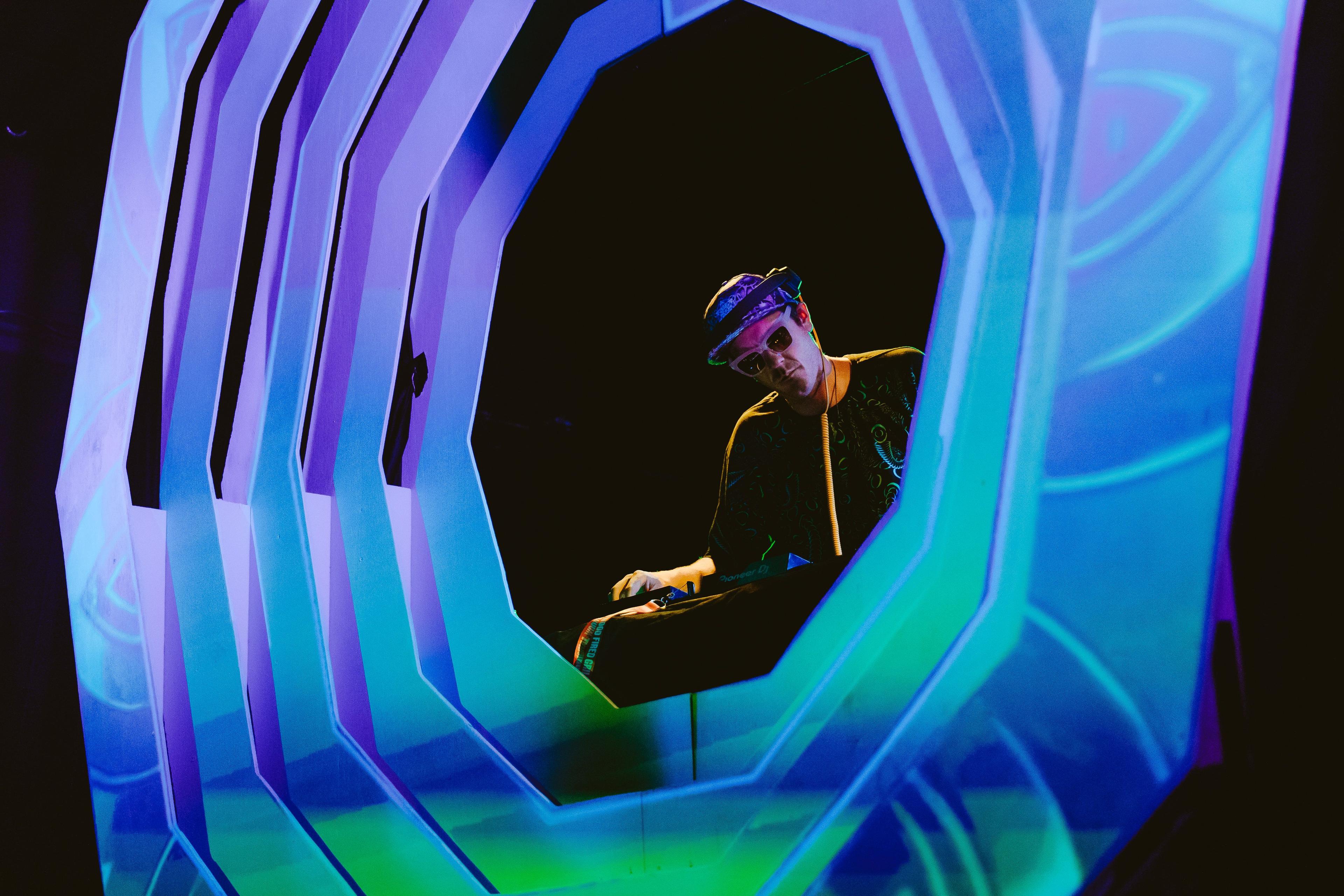 A DJ on-stage, in front of them is a decagonal lighting structure emitting a pattern of purple, green and blue. 