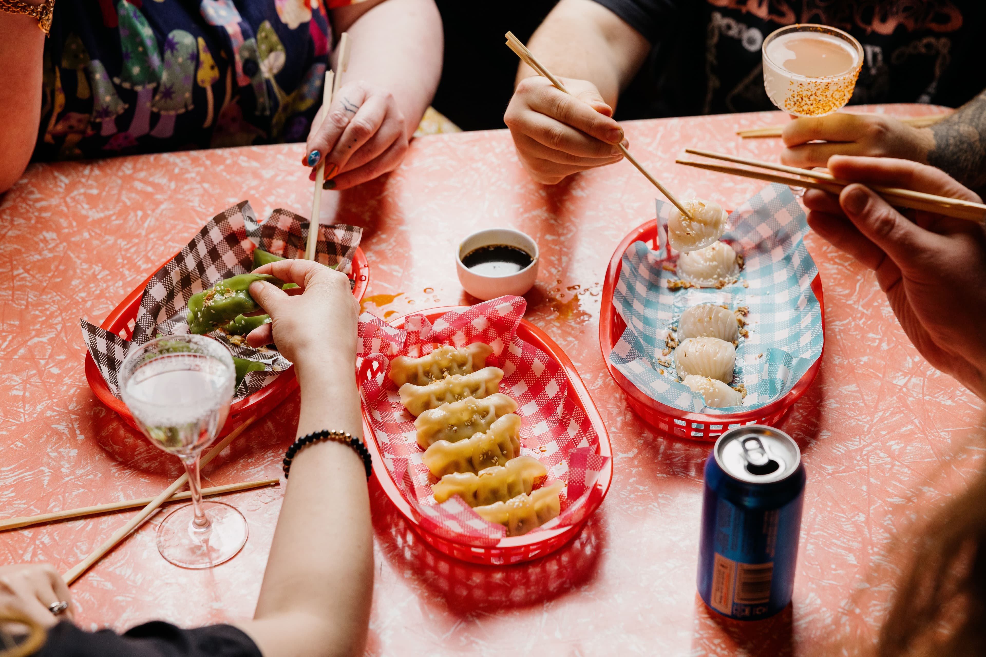 People share dumplings from red baskets lined with checkered paper, using chopsticks. A soy sauce dish and drinks, including cocktails and a beer can, sit on the table.