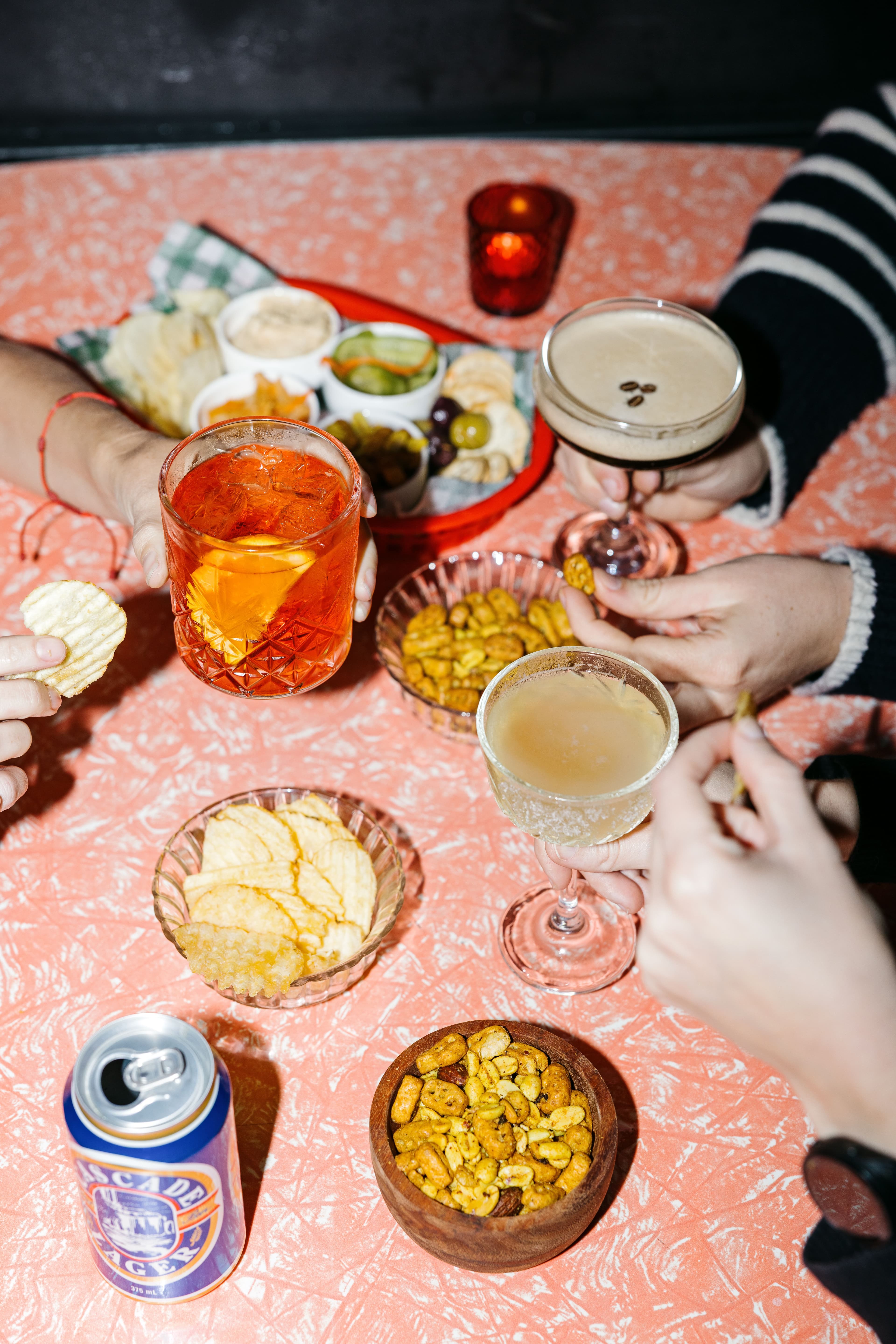 People sharing cocktails and snacks at a retro pink table, with bowls of chips, spiced mix, olives, dips, and a can of Cascade Lager.