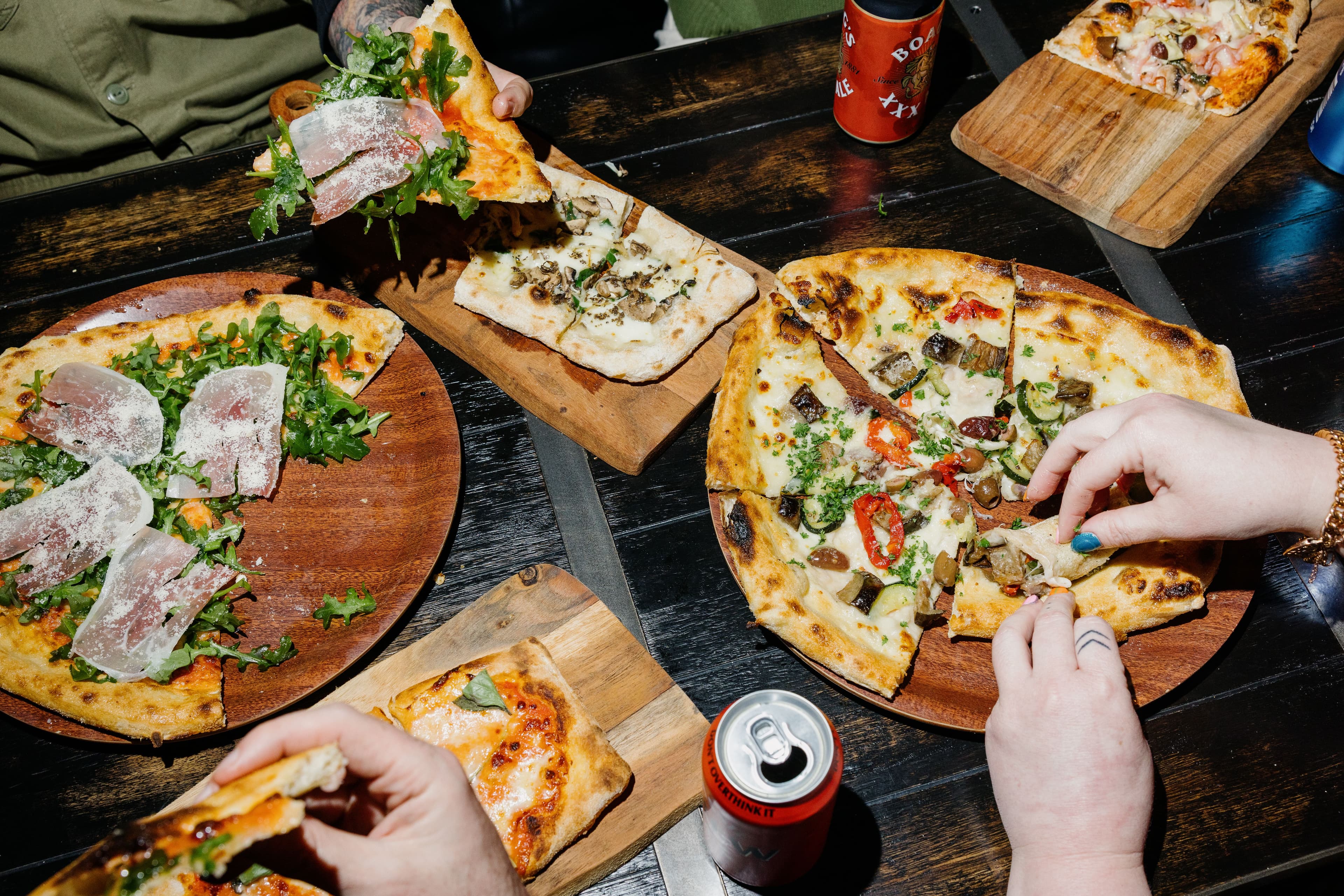 Four people sharing different types of pizza on wooden boards at a dark table, with hands reaching for slices and cans of beer visible.
