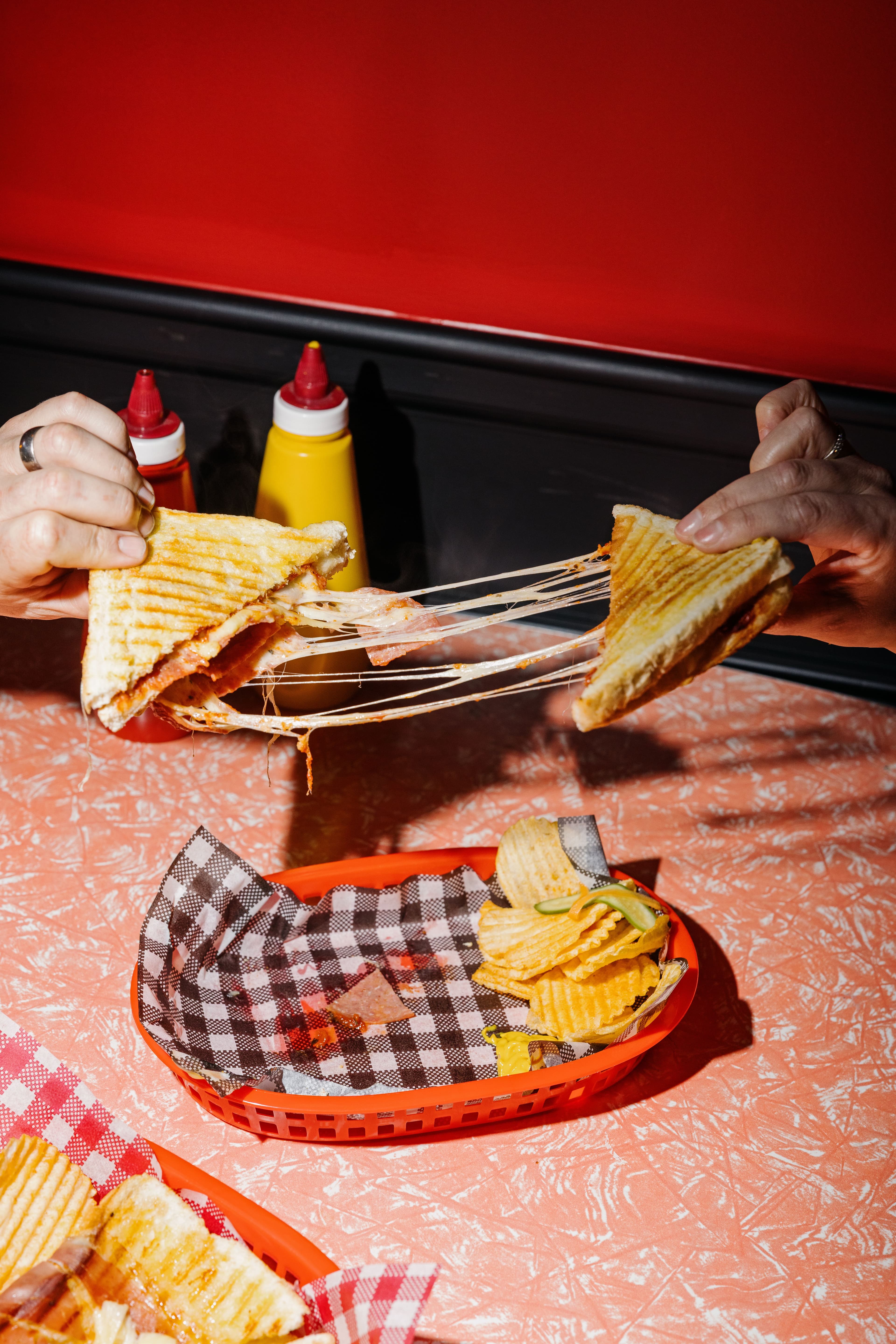 Two hands pull apart a grilled cheese sandwich with melted cheese stretching between the halves; potato chips sit in a basket on a table with ketchup and mustard bottles in the background.