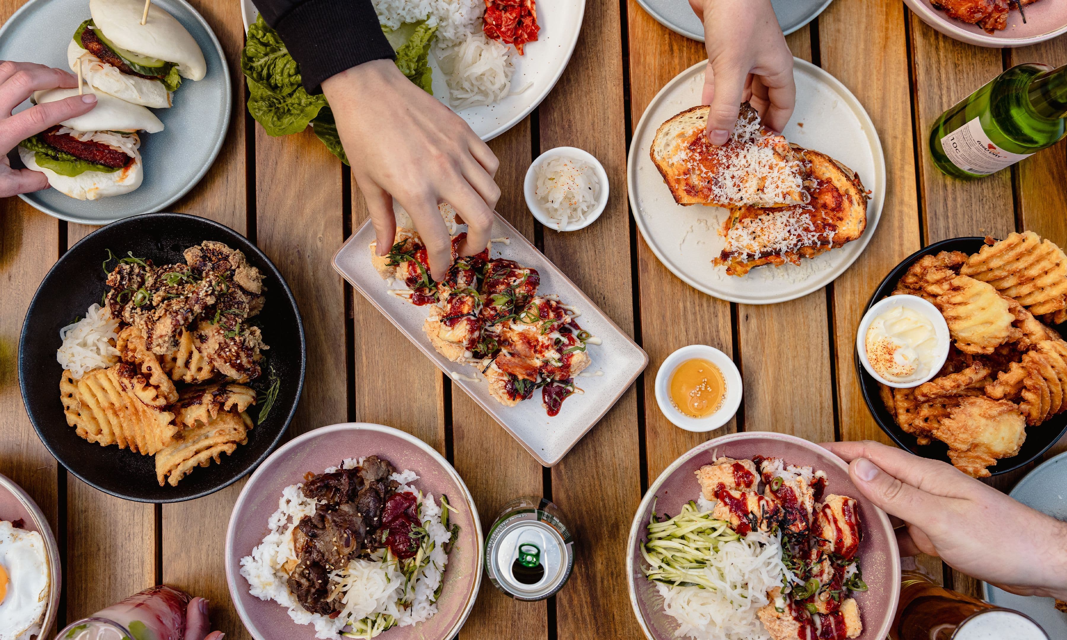 Top-down view of a wooden table filled with a spread of Asian fusion dishes, including bao buns, rice bowls, fried chicken, waffle fries, and small sauce side dishes, as hands reach in to share the meal.
