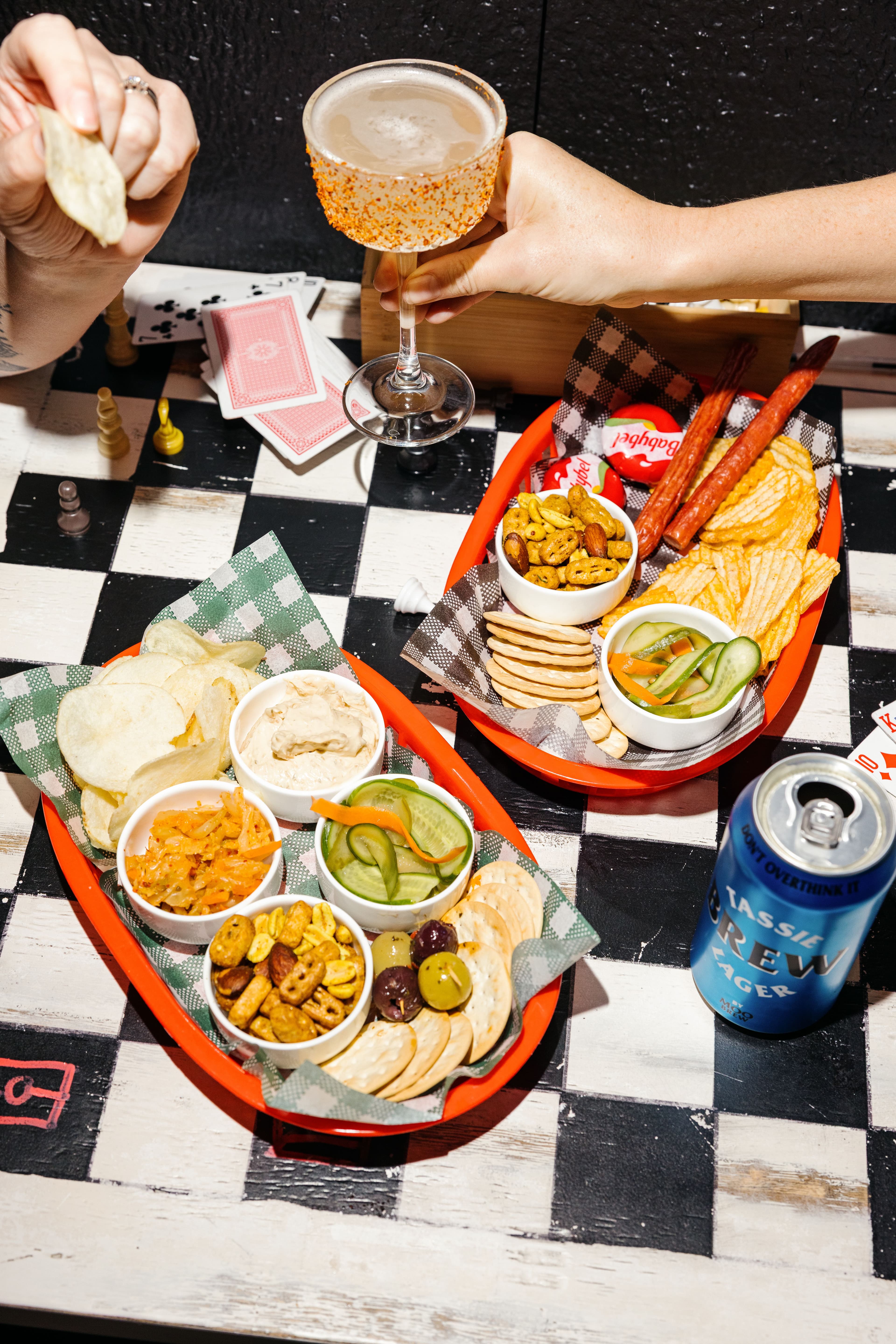 A vibrant spread of snacks and a cocktail on a checkered table, featuring dips, chips, and game pieces in the background.